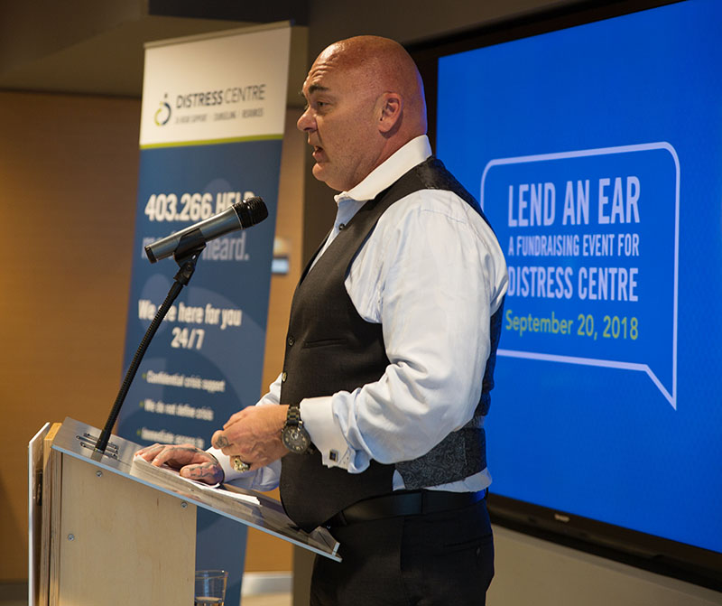 img des: Jim Ryan standing at a podium, speaking at Lend An Ear 2018. There is a Distress Centre banner and a screen behind him showing the Lend An Ear brand - a speech bubble with "Lend An Ear: A Fundraising Event for Distress Centre" written inside. 